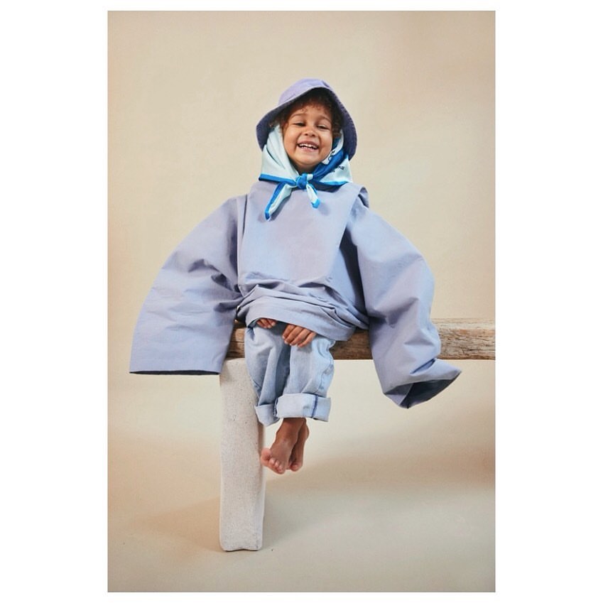 A cheerful child with a toothy grin is seated on a wooden bench. The child is adorned in an oversized periwinkle outfit with a hood.