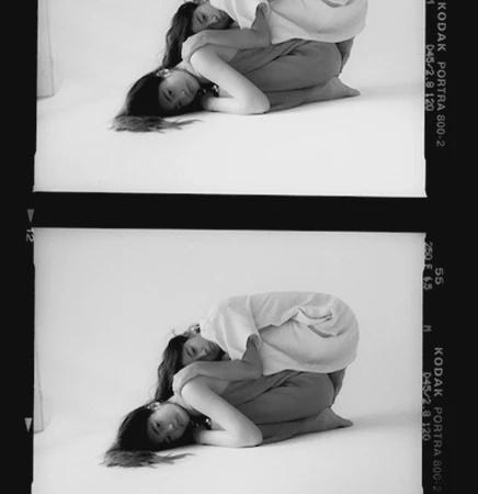 Two duplicate film strip frames showing a woman curled on a white studio floor wearing a light t-shirt and trousers.