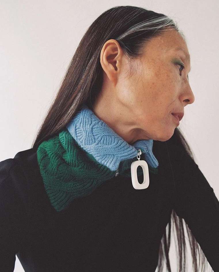Elegant woman with long dark hair featuring grey highlights and wearing a green and blue knitted collared shirt with a white pendant.