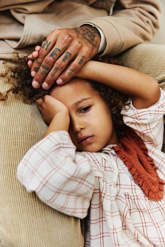 Close-up of a young child lying on a man's lap, resting her head, with his tattooed hand gently on her head in a tender moment.
