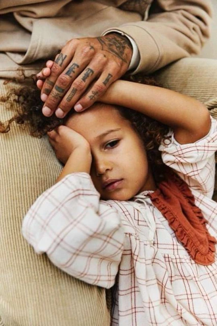 Close-up of a young child lying on a man's lap, resting her head, with his tattooed hand gently on her head in a tender moment.