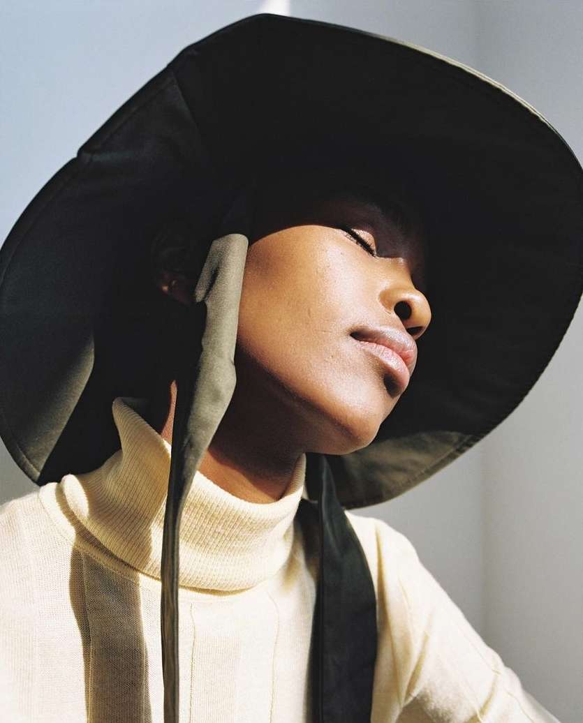 Woman with dark skin poses in cream turtleneck sweater and olive green sun hat, eyes closed, bathed in sunlight indoors.