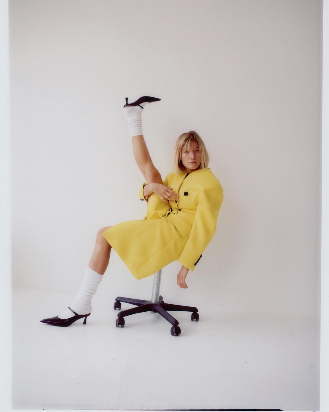 Woman in yellow jacket and white socks sitting on a rolling chair with one leg up. Black heels accent her pose in a clean studio.