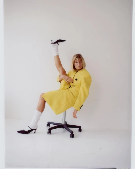 Woman in yellow jacket and white socks sitting on a rolling chair with one leg up. Black heels accent her pose in a clean studio.