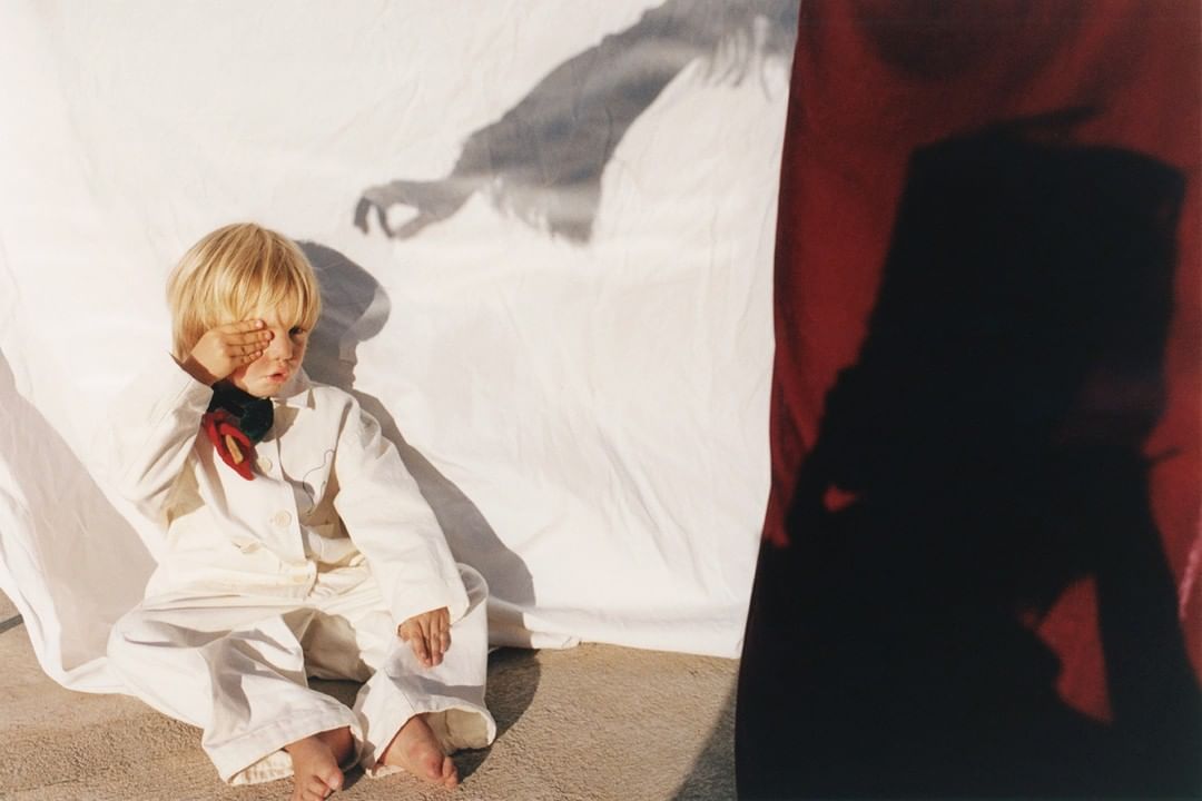 Blond-haired child in a white jumpsuit covers one eye while sitting on the ground with a white sheet and shadows in the background.