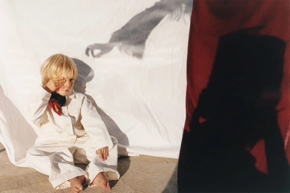 Blond-haired child in a white jumpsuit covers one eye while sitting on the ground with a white sheet and shadows in the background.
