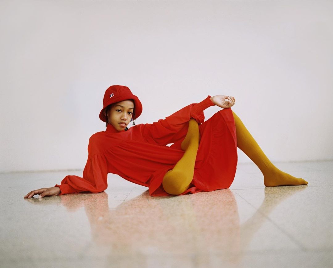 Young girl in a red dress and bucket hat lies on a reflective floor against a white wall. The outfit is complemented with gold tights.