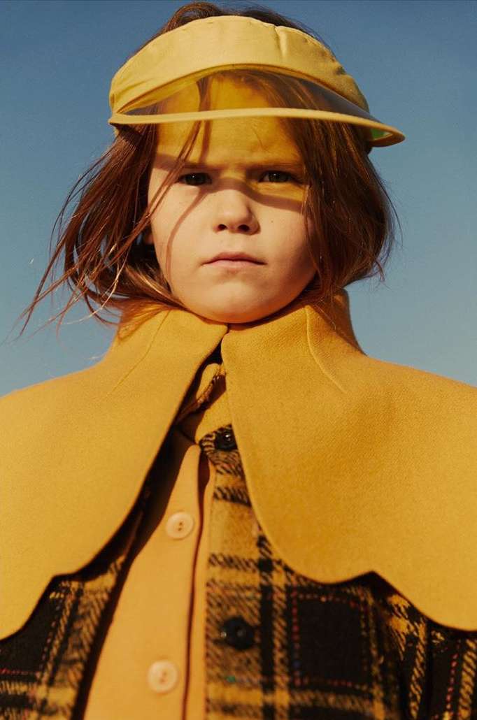 Child in yellow visor and plaid jacket stands outdoors against a light blue sky. Fashion-forward kids apparel for fall.