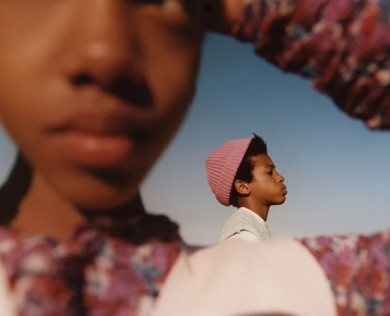 Close-up of two young African-descent children outside under a clear sky. One wears a floral top and the other a pink knit cap.