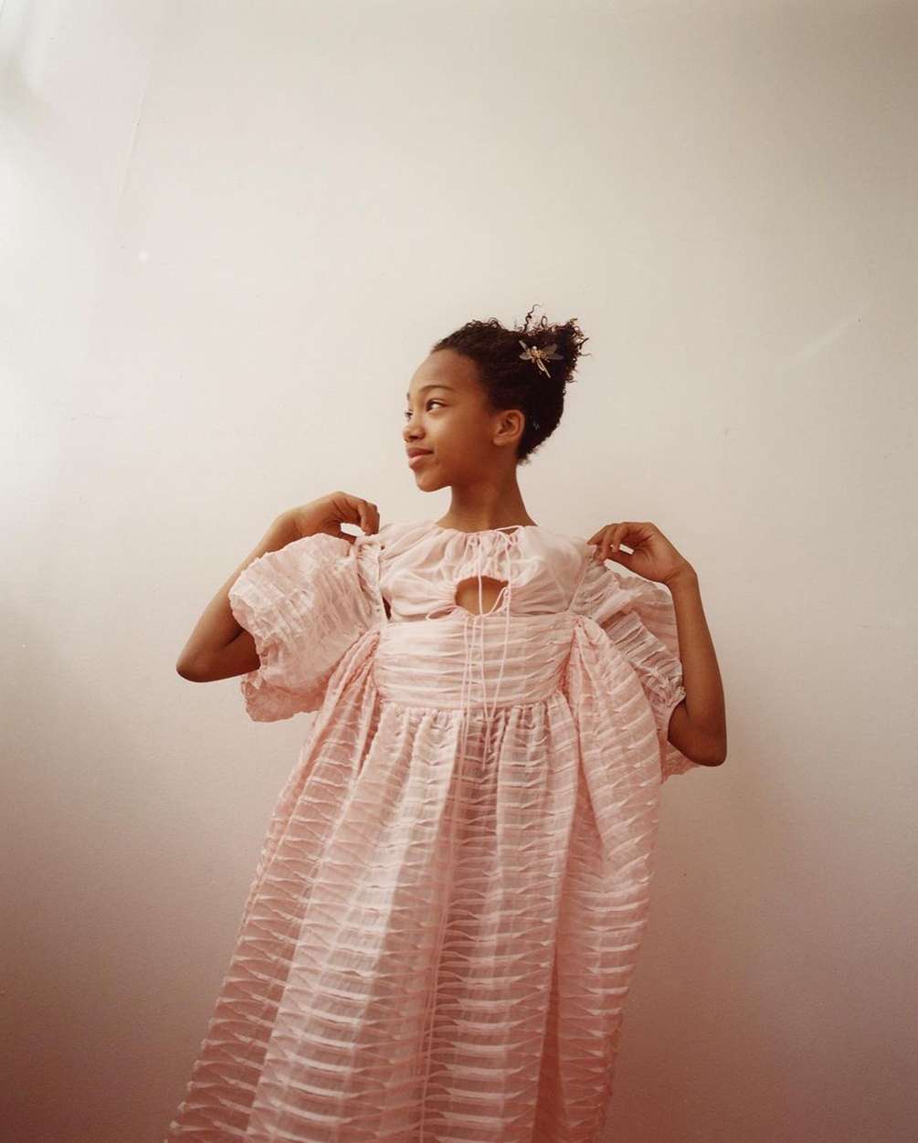 Young girl in a pink, textured dress posing in front of a solid, light pink wall. She is facing to the left, with a slight smile.