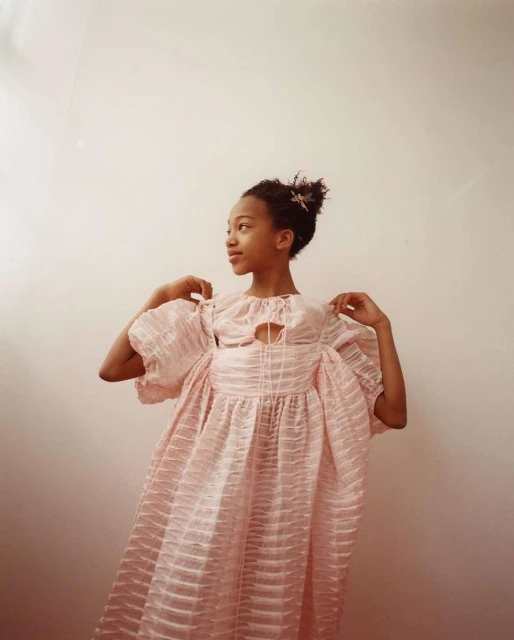 Young girl in a pink, textured dress posing in front of a solid, light pink wall. She is facing to the left, with a slight smile.