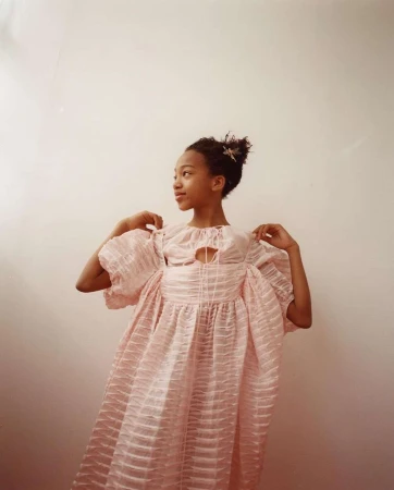 Young girl in a pink, textured dress posing in front of a solid, light pink wall. She is facing to the left, with a slight smile.