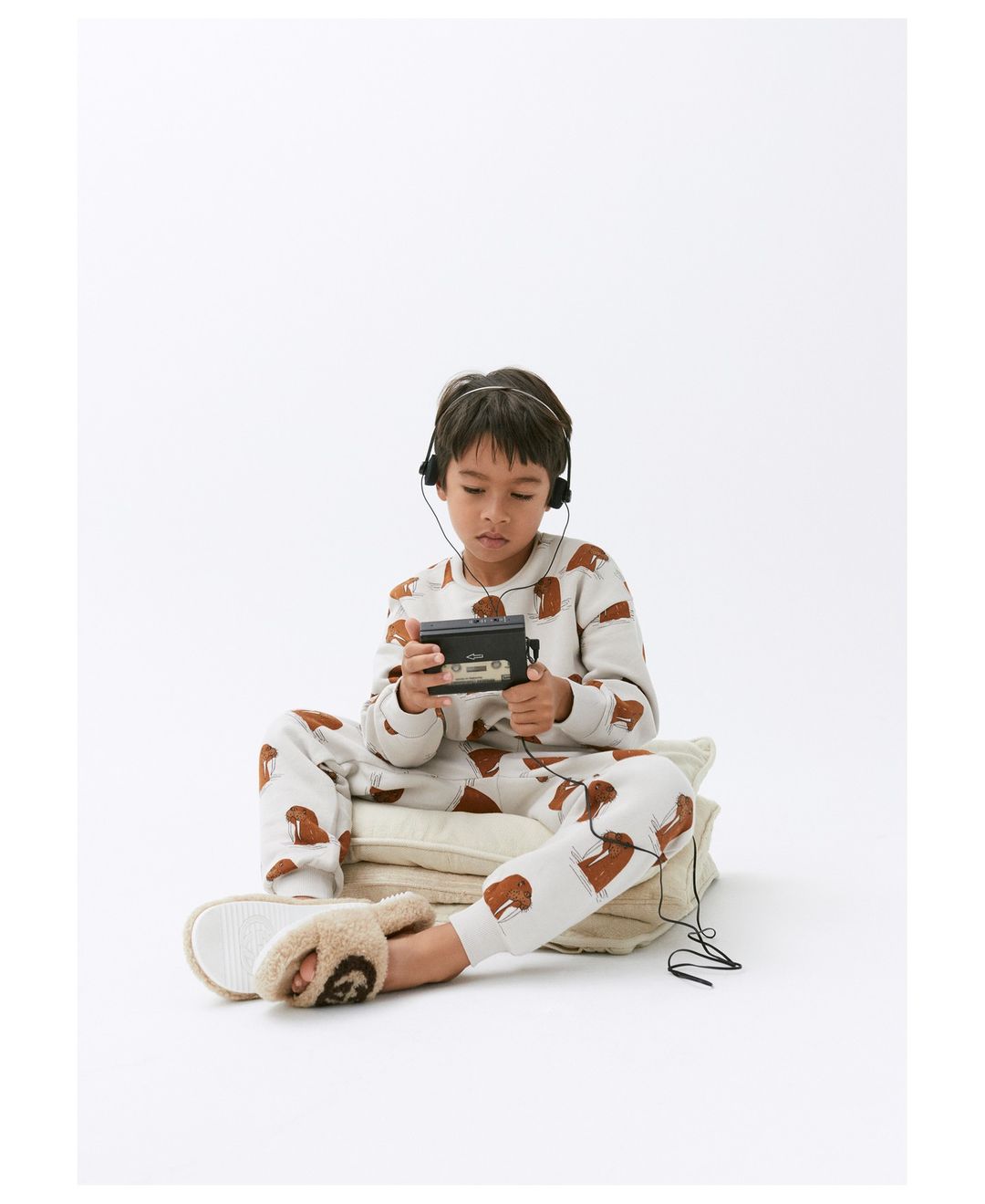 Boy wearing headphones and pajamas with a dog pattern, sits on cushions, holding a cassette player in a bright studio setting.