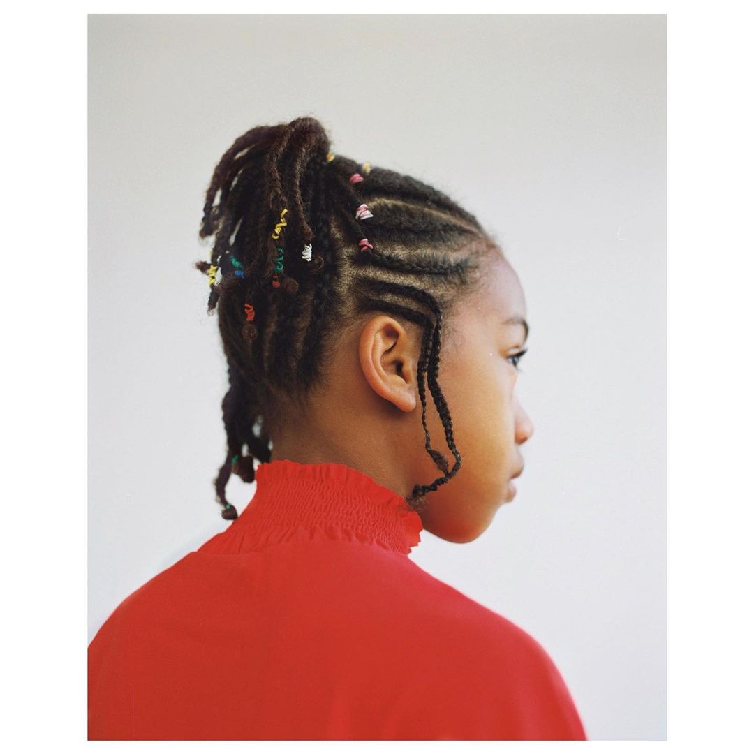 Portrait of a young Black girl in a red top with braided hair and colorful beads, side profile against a white background in studio lighting.