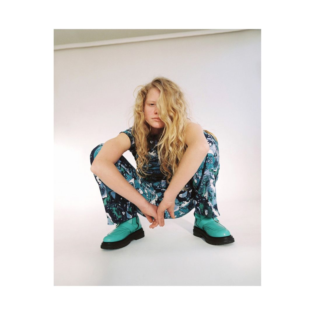 Adult female in a crouching pose, wearing a patterned top and pants with boots, indoors in a photo studio with neutral background.