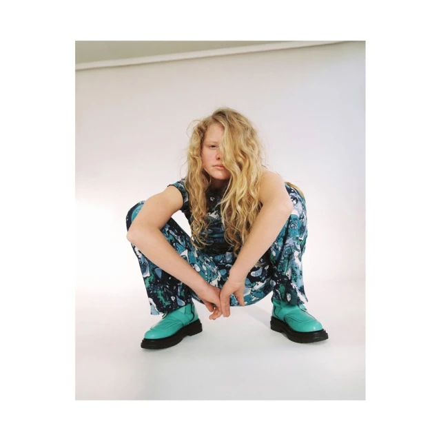 Adult female in a crouching pose, wearing a patterned top and pants with boots, indoors in a photo studio with neutral background.