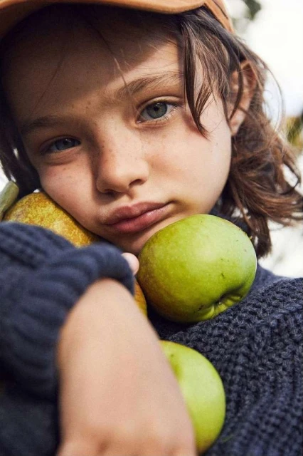 Close-up of child with brown hair and hat hugging green apples and pears, wearing a grey knit sweater, outdoor setting.