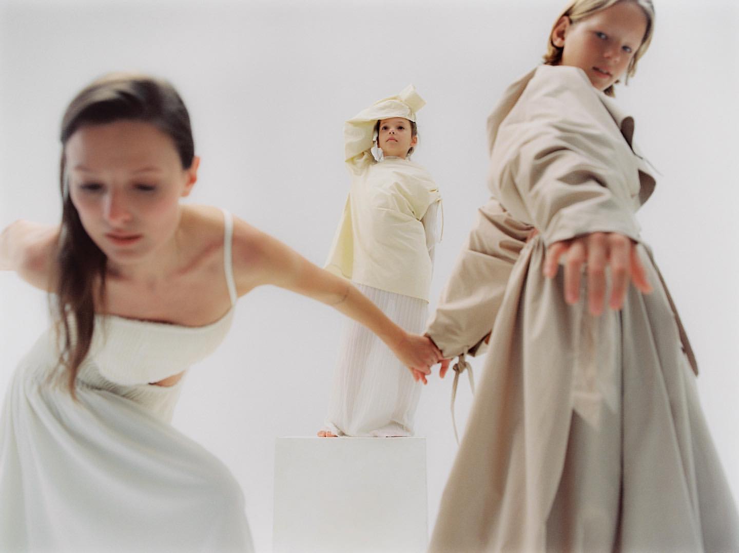 Three fashion models in a minimalist studio setting, wearing light-colored clothing. One stands on a platform, holding hands.