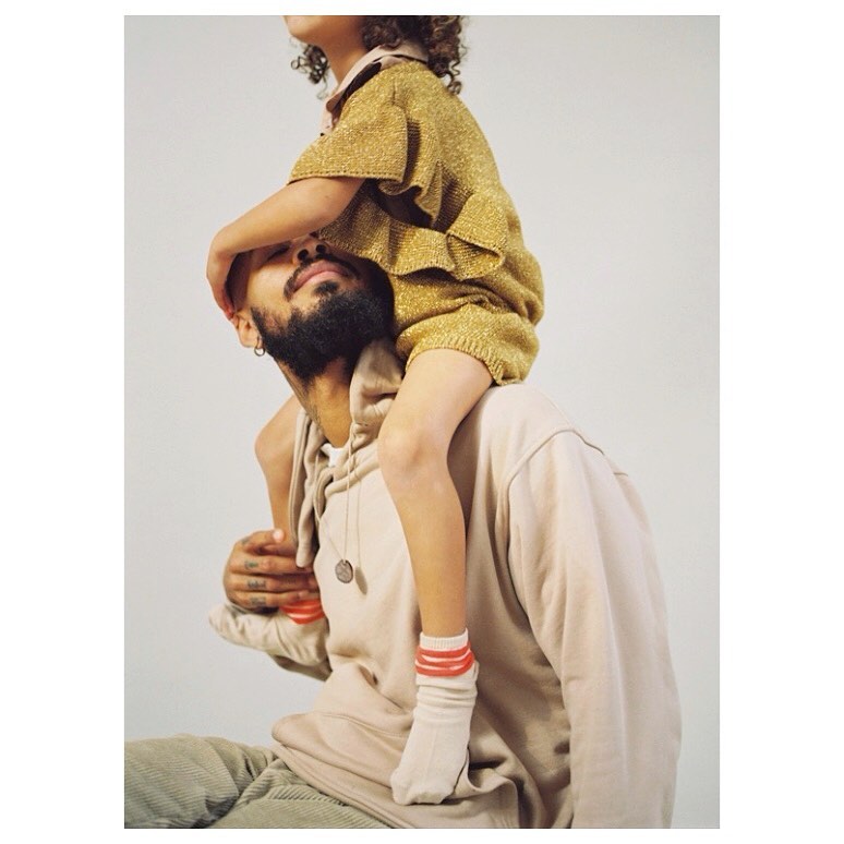 Bearded adult male carries child in a gold outfit on his shoulders in front of a neutral background in a portrait-style studio shot.