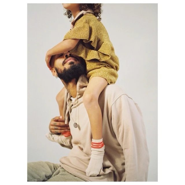 Bearded adult male carries child in a gold outfit on his shoulders in front of a neutral background in a portrait-style studio shot.