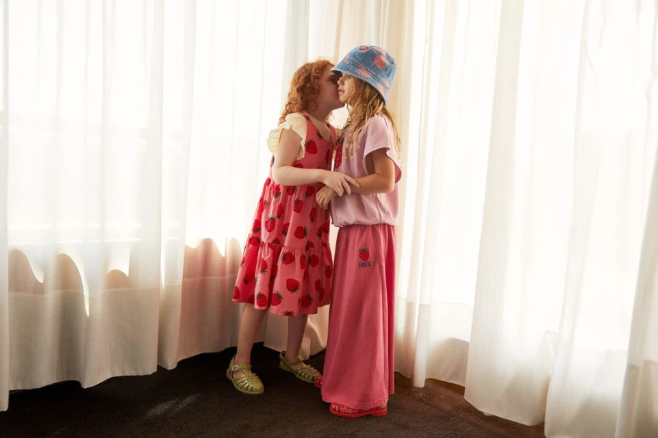 Two young girls embracing and standing by a bright window with sheer curtains. One wears a strawberry dress, the other a pink outfit and hat.