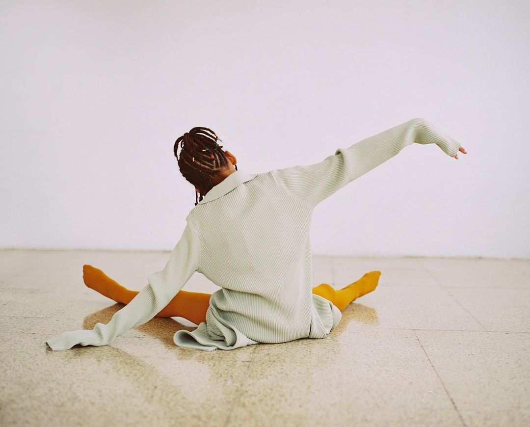 An athletic female adult sits on a studio floor in a light green dress and gold tights, stretching her arms wide. Simple, bright lighting.