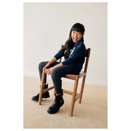 Young child laughing while seated on a wooden chair wearing a dark sweater and black boots against a neutral background.