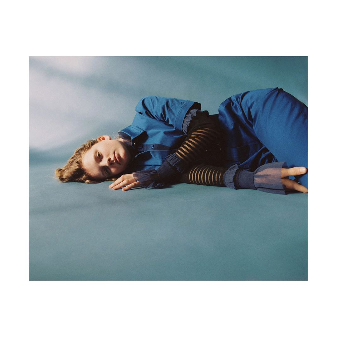 Woman in blue coat and pants lying on a blue surface in a studio environment, with soft, diffused lighting and shallow depth of field.