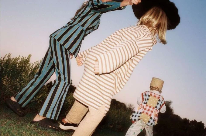 Three kids in retro striped outfits stand on a grassy hill under a bright blue sky, showcasing a vintage fashion editorial.