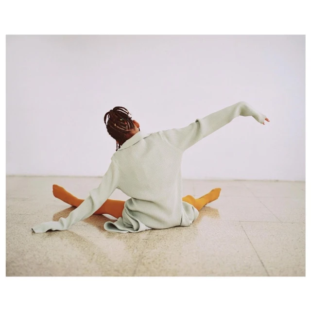 Woman in long-sleeved mint-green shirt and orange tights poses on a light-beige floor against a plain white wall in a wide shot.