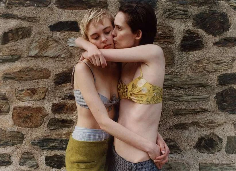 Two adult females embrace affectionately in front of a rustic stone wall, wearing casual summer tops. The scene evokes warmth and intimacy.