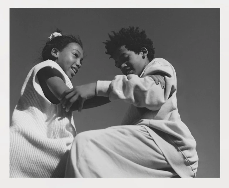 Black and white photograph of two children interacting closely, with a clear sky in the background. Youthful, playful connection captured.
