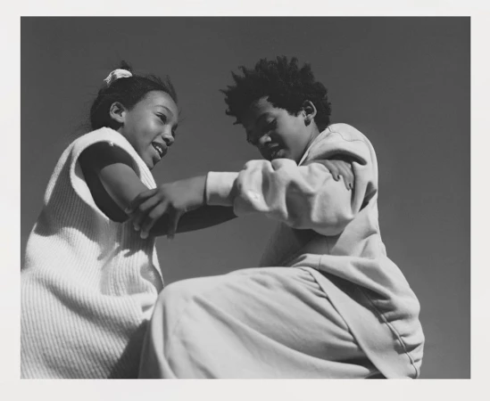 Black and white photograph of two children interacting closely, with a clear sky in the background. Youthful, playful connection captured.