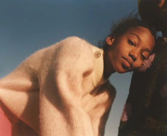 Close-up portrait of a young Black woman in a pink fuzzy sweater leaning against another person under a clear blue sky.