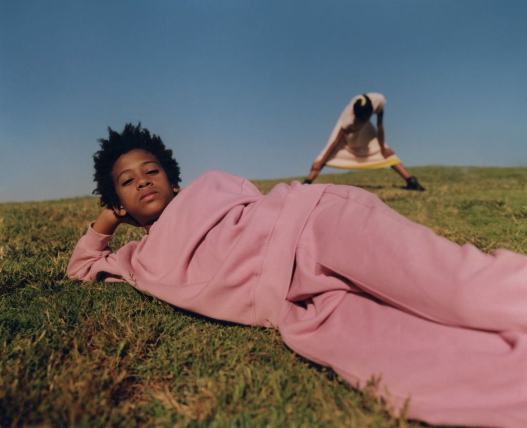 Young person in pink sweatsuit lies on green grass under blue sky; another person stretches in background, outdoors in daylight.