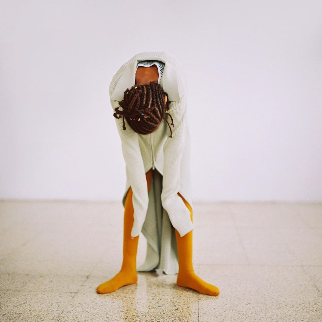 Female figure bending forward in a white robe with yellow leggings against a bright white background, showing modern dance or fashion.