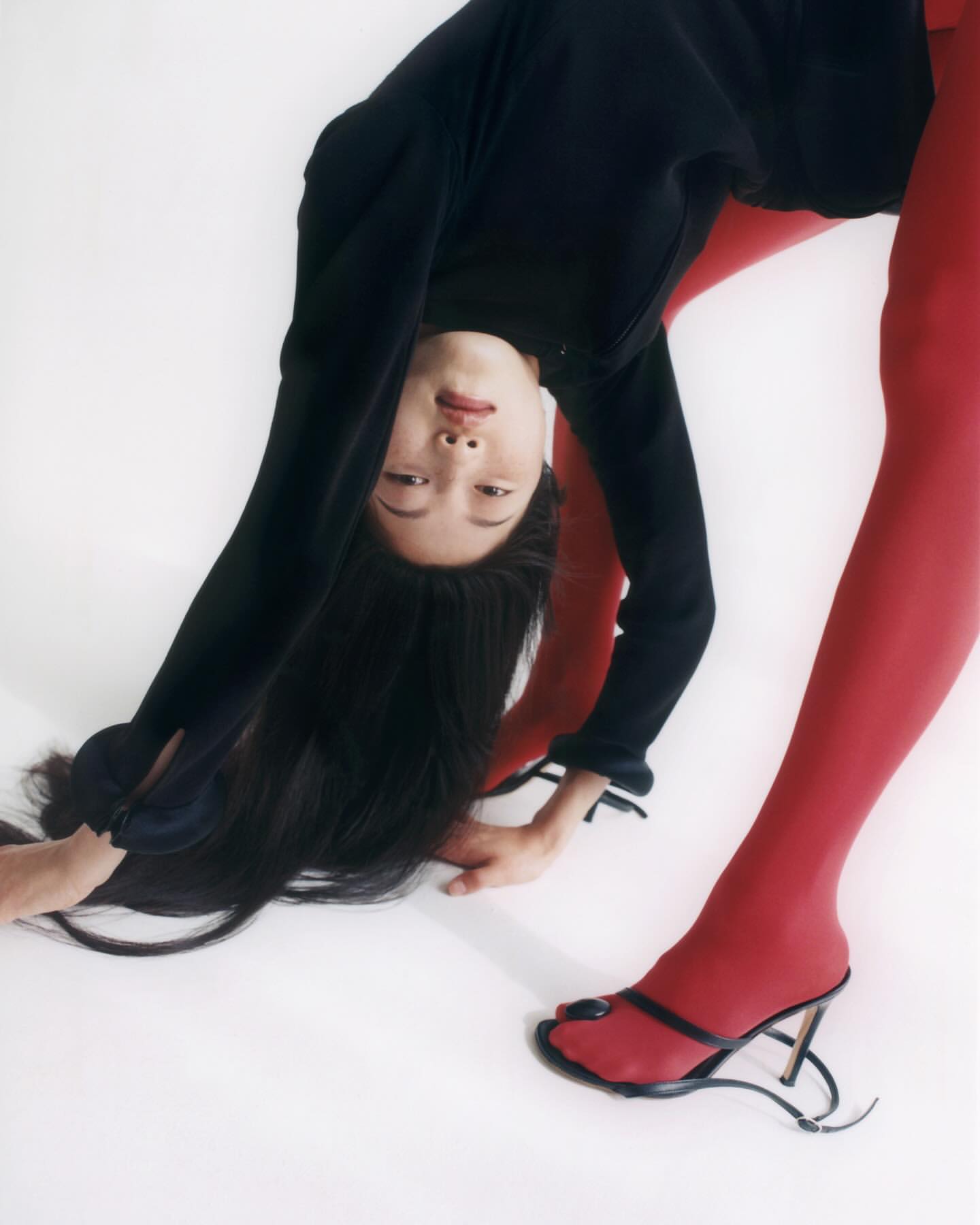 Fashion image of an Asian woman in red tights and black top bending over, facing the camera, wearing black heeled sandals on a white backdrop.