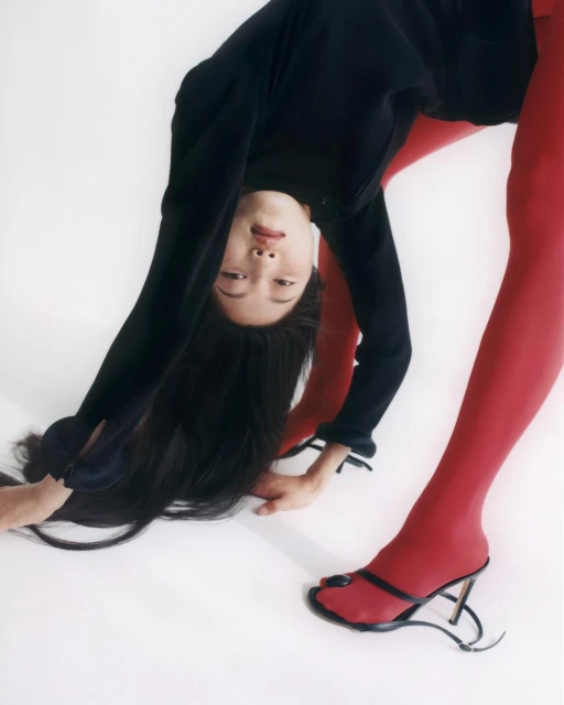 Fashion image of an Asian woman in red tights and black top bending over, facing the camera, wearing black heeled sandals on a white backdrop.