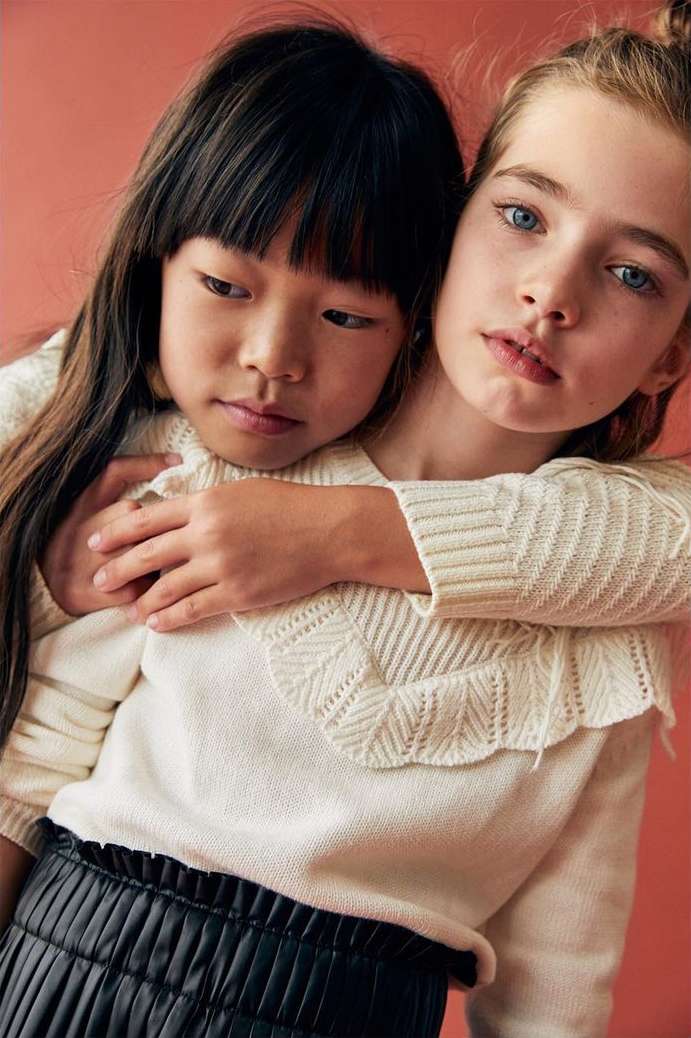 Two young girls, one Asian and one Caucasian, embrace in a close-up shot, wearing matching sweaters, against a pink background.