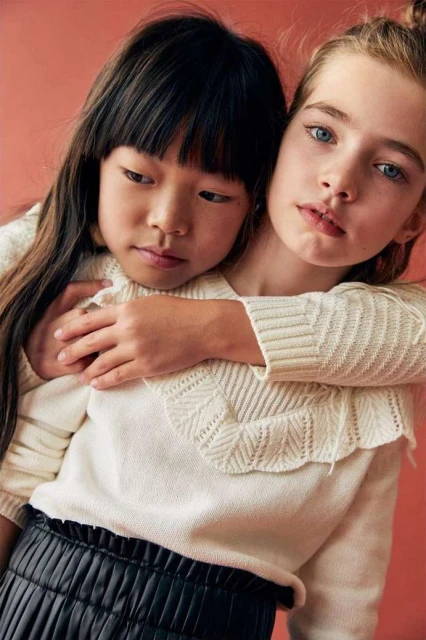 Two young girls, one Asian and one Caucasian, embrace in a close-up shot, wearing matching sweaters, against a pink background.