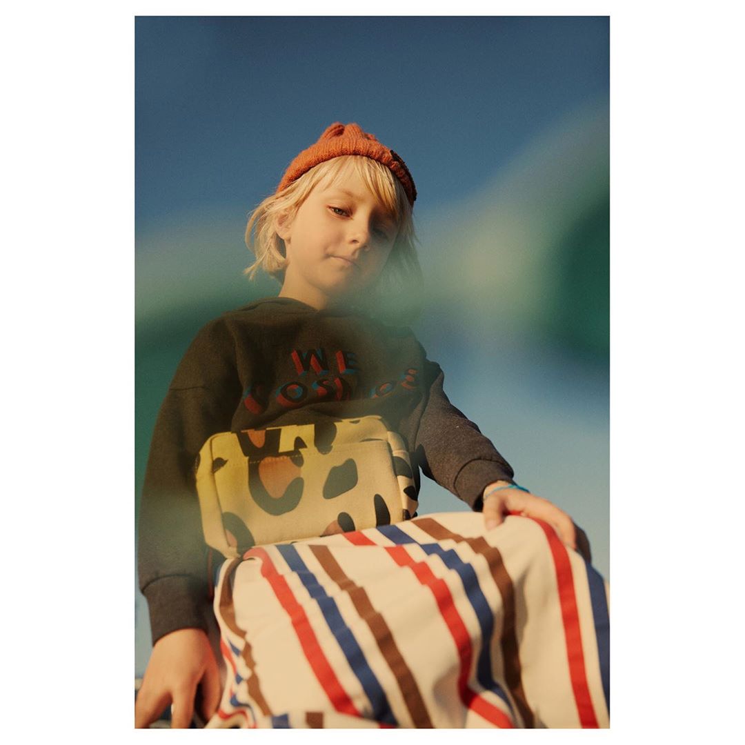 A young child with fair hair wearing a hat and patterned clothing poses outdoors against a blue sky, captured in a portrait.