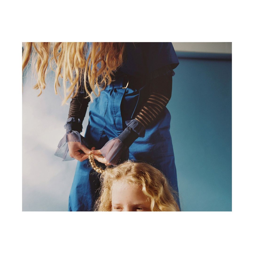A person in blue clothing braids the hair of a blonde child. Shot indoors with a blue background, focus on the hair braiding.