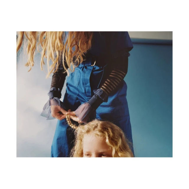 A person in blue clothing braids the hair of a blonde child. Shot indoors with a blue background, focus on the hair braiding.