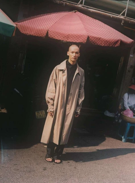 Bald adult stands on city street wearing beige trench coat. Pedestrian umbrella visible overhead. Low angle, natural lighting.
