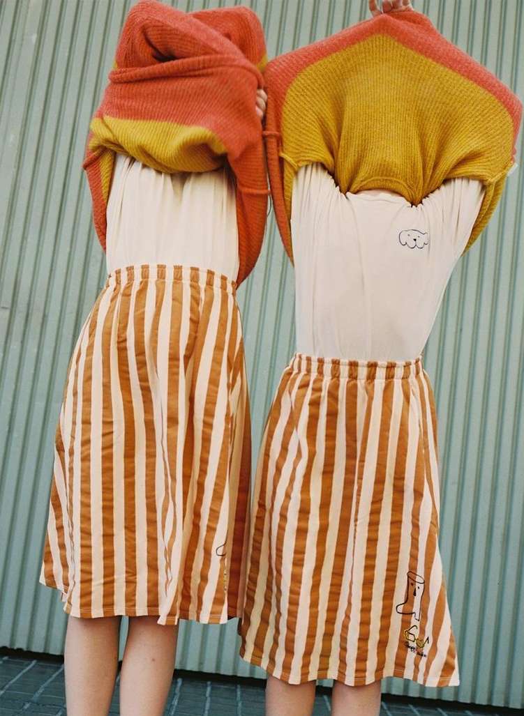 Two individuals showcasing matching striped skirts, posing against a light blue wall, with one person partially removing an orange sweater.