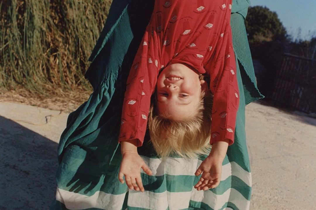A child in a red patterned shirt hangs upside down, supported by an adult in a green and white striped dress outdoors in bright daylight.