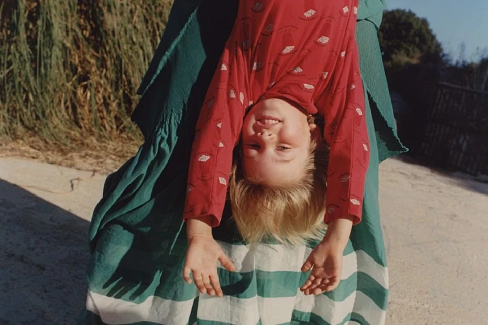 A child in a red patterned shirt hangs upside down, supported by an adult in a green and white striped dress outdoors in bright daylight.