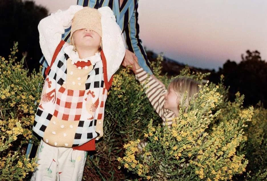 Two children play in a field of yellow wildflowers in the daytime, one covering their eyes, the other partially hidden by foliage.