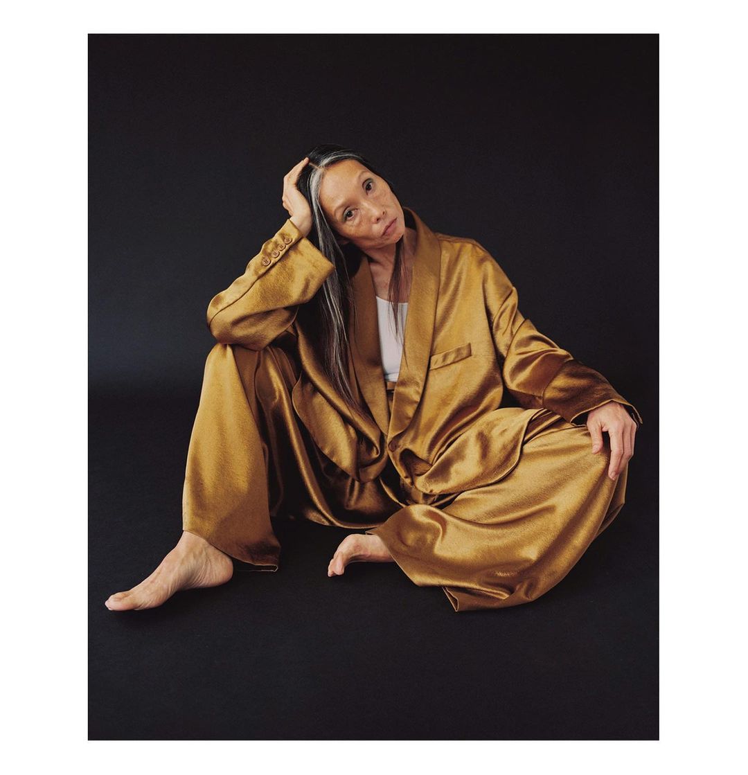 Older woman with gray hair and in a gold silky pyjama set sitting on the floor in a studio, looking at the camera.