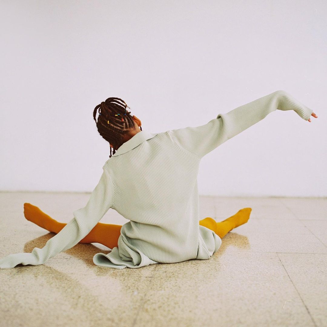 Back view of a young adult woman with braided hair wearing a light green dress and yellow tights sitting on a beige tile floor.